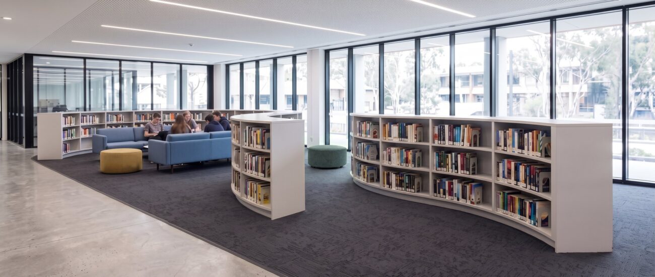 University library with curved double-sided shelving runs, soft seating and full-height glazing